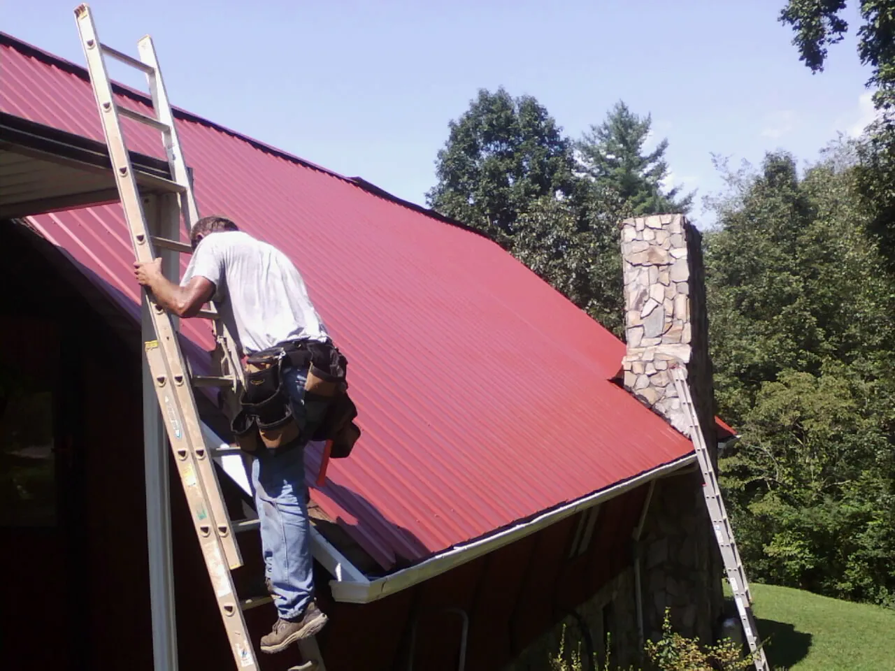 Roofer on a ladder beside a red metal roof and stone chimney — Soffit & Fascia Repair by Roofing Brothers in Pinnacle